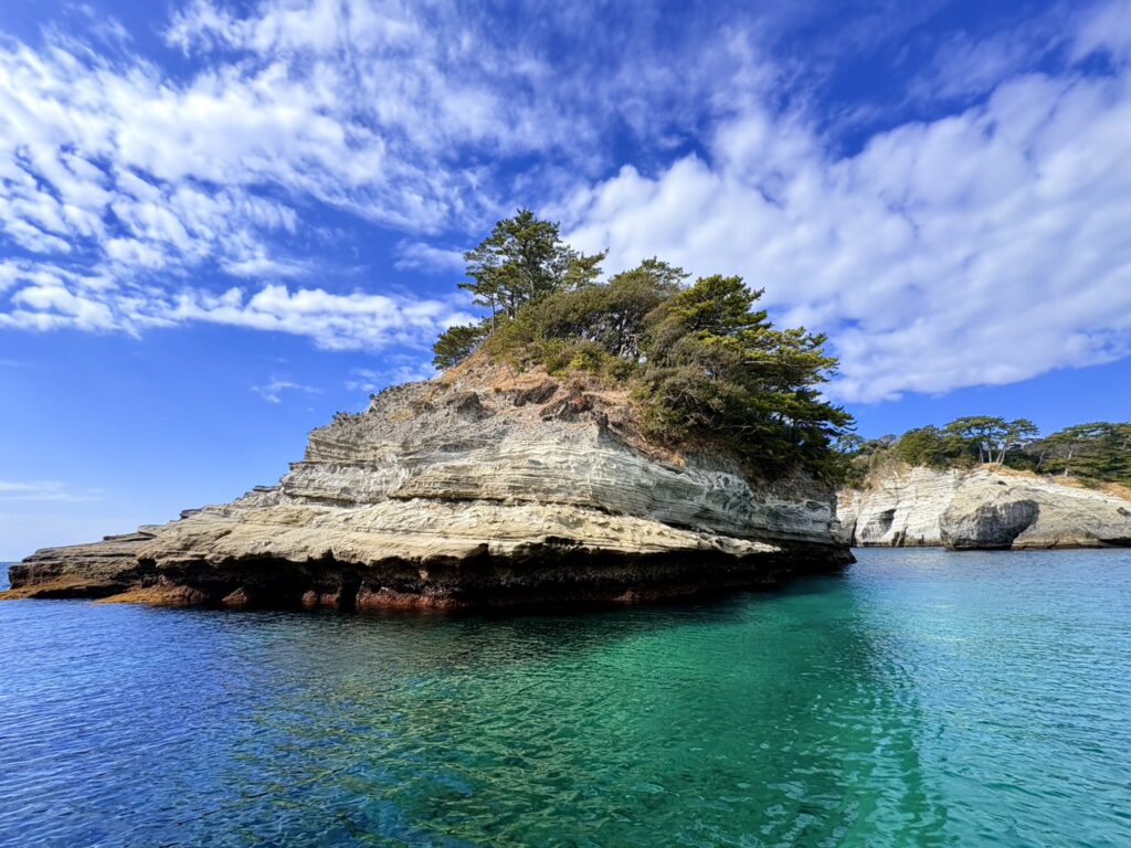 層状の白い岩肌と木がのる小島を、透明度の高い青緑色の海越しに撮影。晴天で青空と雲が広がっている。